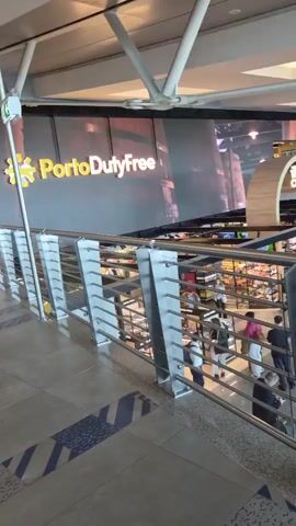 The camera is positioned on an elevated walkway inside an airport, overlooking a lower level of shops and stores. The walkway has a metal railing and the floor has diagonally-oriented black and white stripes. The Porto Duty Free shop is visible directly ahead with a large sign and an electronic display. Passengers are seen walking around and browsing the shops. The ceiling is visible and has white support beams and inset light fixtures. The scene appears to be a typical airport terminal.
