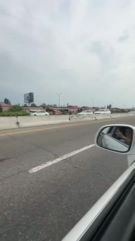 A side-view mirror of a white car shows the reflection of a person holding a phone and a white car driving alongside. The view from inside the car shows a highway with two lanes of traffic separated by a concrete barrier. The sky is overcast. In the distance, there are houses and trees.