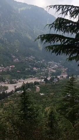 The video shows a panoramic view of a valley with a river running through it. Buildings are scattered on the hillsides, and trees cover the slopes. The sky is overcast, and the overall mood is serene and peaceful. The camera slowly pans across the landscape, offering a breathtaking vista of nature and human settlement.