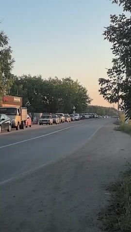 The video shows a traffic jam on a road with trees and buildings lining the side. Several cars are stopped in a line, with some people standing near them. A small building with a sign that says "КОФЕ" (coffee) is visible in the background. The sky is clear and the sun is shining.