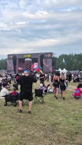 A wide shot captures a lively outdoor concert scene under a cloudy sky. A large stage, adorned with the word "YLETAY" and illuminated screens, stands in the background. Several people are visible on stage, and large screens display graphics and possibly live footage of performers. In the foreground, a diverse crowd of people is spread out across a grassy field, many seated in camping chairs or on blankets. Some individuals are standing and appear to be watching the performance. The atmosphere suggests a festival or large public gathering. The camera is positioned behind a few people in the foreground, including a person in a black t-shirt and shorts, facing the stage. The overall environment is a wide-open space with trees visible in the distance behind the stage and tents or other structures on the far right.
