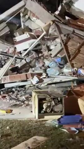 The video shows the wreckage of a collapsed building at night. The pile consists of bricks, concrete, lumber, furniture, and household items. A generator is visible at the base of the pile. There is grass on the ground and a streetlight in the distance.