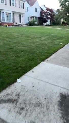 A person is jogging on a sidewalk. The person is wearing a dark t-shirt, light-colored shorts, and a hat. The jogger is on the right side of the frame, moving away from the camera. A green lawn is to the left of the sidewalk, with houses visible in the background. Trees line the street on the right side of the sidewalk. The scene appears to be a suburban neighborhood during the day.