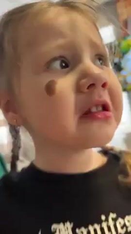 A close-up video shows a young girl with blonde hair in pigtails, wearing a black shirt, looking upwards with a concerned expression. She has a noticeable mole on her left cheek. The background is slightly blurred but suggests an indoor setting. She speaks, and her words are "дует" (blows) and "может мне на глазку водой" (maybe on my eye with water).