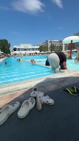 The video shows a resort swimming pool on a sunny day. The pool is filled with bright blue water, and there are several people swimming and relaxing. In the foreground, a collection of white and purple Crocs and a pair of yellow and black flip-flops are scattered on the poolside mat. A woman in a black top and light-colored pants is bending down by the edge of the pool, reaching into the water. A man in dark swim trunks is standing in the pool, facing away from the camera. Another man, also in swim trunks, walks out of the pool, holding a dark object. In the background, resort buildings and palm trees are visible, and other people are scattered around the pool area. The sky is clear and blue with some white clouds.