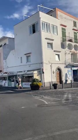 The video opens with a view of a white building with multiple windows and balconies in a European town. A person walks by the building. The camera pans to the right, showing more buildings with shops and balconies. The scene transitions to a street with a cobblestone pavement and a pedestrian crossing. A motorcycle and a car are visible on the street, along with a domed building in the background. The video then shows a person wearing a hat and a dress, standing on a sidewalk next to a street with parked motorcycles and scooters. The scene changes to a harbor with several boats docked and flags flying, including the Italian flag. The video ends with a view of the harbor and boats under a clear blue sky.