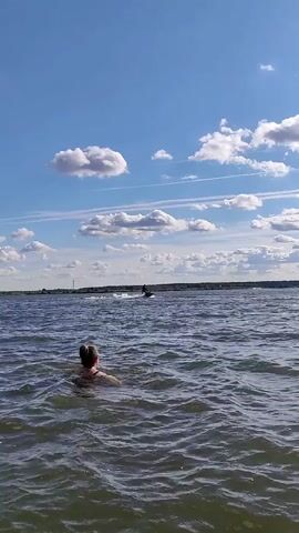 A person is swimming in choppy water under a bright blue sky with scattered white clouds. In the distance, a jet ski is visible, creating a spray of water as it moves across the lake. The shoreline is lined with trees and a tall tower can be seen in the distance.