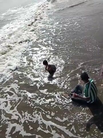 A wide shot captures a sandy beach where two individuals are interacting with the ocean's edge. In the center of the frame, a young person is partially submerged in the water, appearing to be playing or splashing. To the right, an adult is seated on the sand, close to the water line, also engaged with the scene. The ocean is active, with waves breaking and creating foam that reaches the shore. The lighting suggests it is daytime, with sunlight reflecting off the water's surface. The overall environment is a coastal setting.