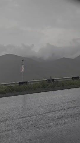 The video shows a grey, overcast day with rain falling on a wet asphalt road. In the background, mountains are visible, partially obscured by clouds. A guardrail runs along the side of the road, with a warning sign indicating a curve to the right. The overall atmosphere is gloomy and damp.
