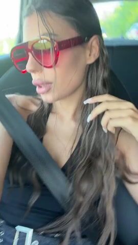 A young woman wearing a black tank top and denim shorts with a silver belt is sitting in the driver's seat of a car. She has long, dark, wavy hair and is wearing large red sunglasses. She is looking towards the camera and her mouth is open as if she is speaking or singing. A black seatbelt is across her chest. The interior of the car is visible, including the car's seat and part of the dashboard. The background outside the car appears to be green foliage.