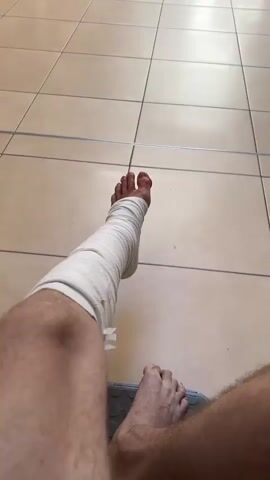 The media file shows a first-person perspective of a person's bandaged leg and foot, resting on a tiled floor. The floor tiles are a light beige color with dark grout lines, and the overall lighting suggests an indoor environment. The leg is wrapped in a white elastic bandage from the ankle to just below the knee. The toes of the foot are visible, with one toe appearing red, possibly due to injury or irritation.

In the background, the scene is a long, well-lit corridor or waiting area. The floor continues with the same beige tiles. Several people are visible in the distance, sitting on benches or standing. One person is wearing a white t-shirt and shorts and appears to be looking down at their phone. Another person is wearing a grey and white patterned top and dark pants, walking away from the camera. Further down the corridor, a man is sitting in a wheelchair, and another person is standing near him. There is a poster or sign on a pillar in the background. A Coca-Cola vending machine is visible on the left side of the corridor. The overall atmosphere appears calm and somewhat sterile, possibly a hospital or clinic waiting area. The presence of the bandaged leg suggests an injury or post-operative condition, and the waiting individuals imply a medical setting. The time of day is not explicitly determinable, but the bright lighting suggests daytime. There is no indication of weather conditions. No specific location cues other than the tiled floor and generic interior features are present within the image itself. The visible text includes the Coca-Cola logo on the vending machine and some text on a sign in the background which is illegible.