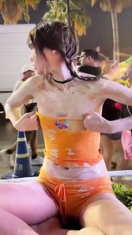A person is sitting down, covered in a white substance, possibly paint or mud. They are wearing an orange tube top and orange shorts. Their hands are pulling at the top of their tube top. In the background, palm trees are visible, and there are other people and some market stalls. The lighting suggests it is nighttime.