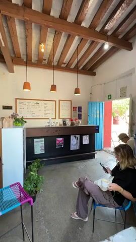 The interior of a café is captured in a medium shot. A woman with blonde hair is seated on a blue chair, holding a white mug and using her phone. She is dressed in a black shirt and brown pants. In the background, another person wearing a white hat is visible near an open door, which is painted turquoise on the inside and red on the outside. To the left, there is a small table with a blue and pink woven chair. Plants are placed next to the table and behind the chair. A service counter runs across the width of the room, with black panels displaying posters. Above the counter, there are two framed whiteboards with handwritten menus. Three pendant lights hang from the wooden beamed ceiling. The floor is concrete, and the walls are painted white.