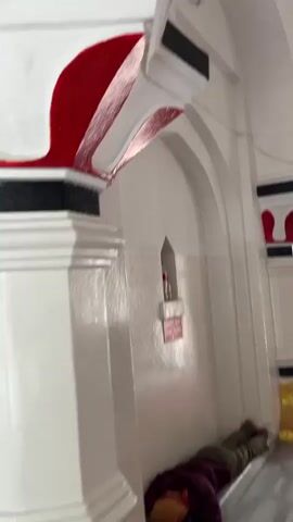 The video shows the interior of a mosque. A person lies on the floor in the foreground. The camera pans to show the dome of the mosque, which has decorative elements. Several people are seen sitting or kneeling on prayer rugs. An arched doorway leads to an outdoor area.