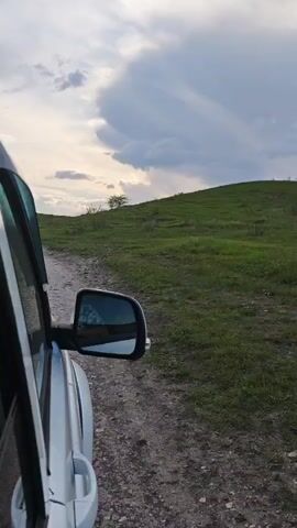 The view from inside a car driving on a dirt road showcases a landscape of rolling green hills against a sky filled with dramatic clouds. The car's side mirror is visible in the foreground, reflecting the surrounding environment. The road appears uneven and rugged, suggesting off-road travel. The clouds in the sky are a mix of bright whites and darker grays, indicating varying weather conditions. The overall scene portrays a sense of adventure and exploration in a natural setting.