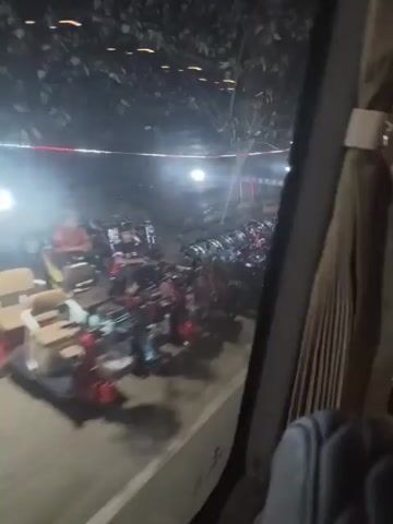 A dimly lit outdoor scene at night, viewed from inside a vehicle. In the foreground, reflections on the window obscure some of the view. Beyond the window, a large number of motorcycles are parked in rows. Some motorcycles have red and black colors, while others are blue and black. Beyond the motorcycles, there are more lights and structures, including what appears to be an arched entryway with decorations. The overall impression is of a busy gathering or event at night, with many people likely present, though most are not clearly visible due to the distance and lighting conditions. The video also captures the side of the vehicle, including a gray seat and the textured window frame.