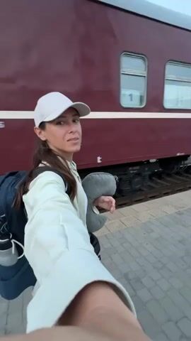 A woman wearing a white baseball cap and a white jacket stands on a train platform next to a maroon train. She has a backpack on and is holding a gray neck pillow. She is looking at the camera.