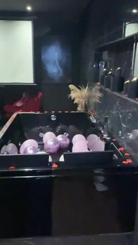 A video tour of a room decorated for a romantic or sensual experience. The room contains a large black bathtub filled with pink and white balloons, rose petals, and what appear to be small decorative balls. There are also several lit candles scattered around the room, along with rose petals on the floor. A massage table, a large screen projector, two red chairs, and a bed with dark grey linens and pillows are also visible. The overall ambiance is dark with accent lighting.