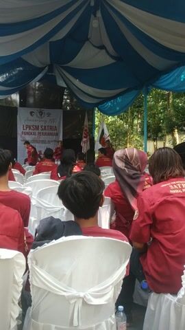 The video shows a group of people seated in white chairs under a blue and white striped canopy. They are facing a stage with a banner that reads "LPKSM SATRIA PANGKAL PERJUANGAN". Most of the people are wearing red shirts with "LEMBAGA PERLINDUNGAN KONSUMEN SATRIA PANGKAL PERJUANGAN" printed on the back. Some individuals are wearing hijabs. The environment seems to be an outdoor setting with trees visible in the background.
