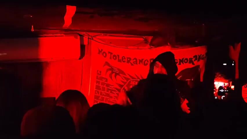 A person wearing a hooded jacket is performing on a stage bathed in red light. Behind them is a banner with the words "NO TOLERANCE FOR IGNORANCE" and an image that includes what looks like a dragon or monster clawing at a brick wall. Several people are in the audience, and some are holding up their phones to record the performance. The scene is dark, adding to the intensity of the performance.