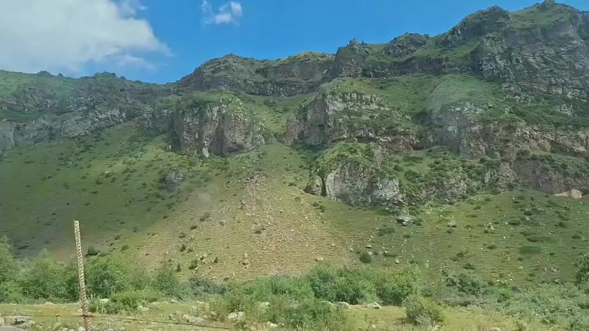 The video shows a time-lapse view of a green, rocky mountain range under a bright blue sky with scattered clouds. The sun shines down, casting shadows across the landscape. Vegetation, including trees and shrubs, covers the lower slopes of the mountains, transitioning to sparse grass and rocks higher up. The video pans slowly to the right, revealing more of the mountainous terrain. No people or specific actions are visible.