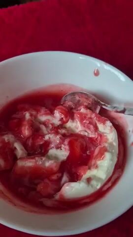 A close-up shot shows a white bowl filled with vanilla ice cream and strawberry topping. A silver spoon is actively mixing the ice cream and topping, creating swirls of red and white. The bowl sits on a textured red surface, possibly a tablecloth or fabric. The lighting is dim, with a slight glare on the spoon and ice cream. The action suggests someone is preparing to eat the ice cream.