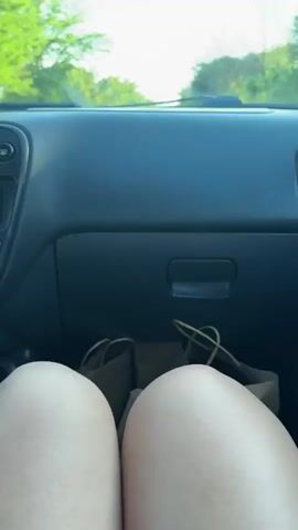 The video shows a first-person perspective from inside a car, looking out the windshield. The dashboard of the car is visible in the foreground, with a phone and some bags on the passenger side. The road ahead is straight and lined with trees. The sky is bright and appears to be daytime. The car is moving forward.