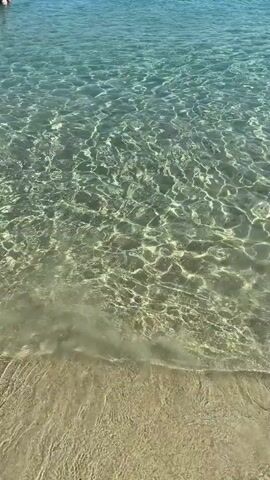 A first-person perspective video shows clear, shallow ocean water with sunlight reflecting off the surface. The bottom is sandy and visible through the water. Gentle waves lap at the shore, creating ripples. The overall scene is peaceful and serene.