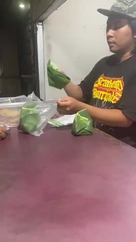 A person wearing a black t-shirt with a graphic design and a camouflage baseball cap is standing behind a maroon counter. They are holding a clear plastic bag in their left hand and appear to be placing an item wrapped in green leaves into it with their right hand. Another similar item, also wrapped in green leaves, is visible on the counter in front of them. A clear container is to the left of the person, and a portion of a pink chair is visible on the far right. The background is dimly lit and appears to be an indoor setting.