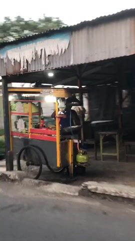 The video shows a food cart at dusk. The cart is yellow with red trim and has a metal roof. There are lights on the cart, illuminating the food being prepared. A propane tank is visible at the back of the cart. A person is partially visible behind the cart, seemingly preparing food. The cart is parked on a street next to a building with a corrugated metal roof.
