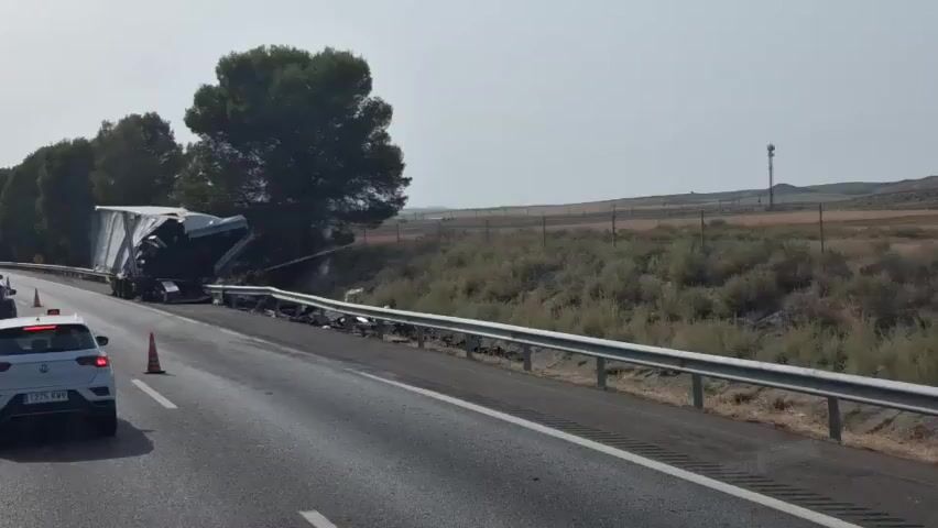 A semi-trailer truck has crashed and overturned on the side of a highway. The trailer is severely damaged, with its contents spilled onto the road and shoulder. Debris from the crash is scattered along the guardrail. A white car is visible in the foreground, stopped on the highway. Traffic cones are placed on the road, and police tape is visible in the background, indicating the area is secured. The landscape appears arid, with sparse vegetation and distant hills under a clear sky.