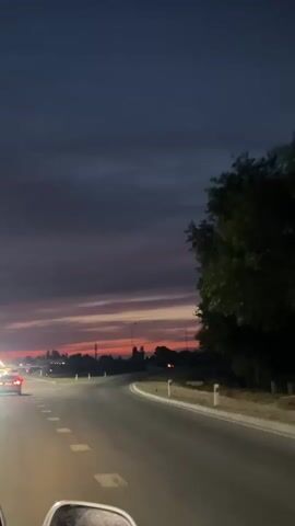 The video shows a view from a vehicle driving on a road at dusk. The sky is a gradient of dark blue and purple, with streaks of pink and orange from the setting sun. Silhouettes of trees and distant buildings can be seen against the horizon. Streetlights and car headlights illuminate the road ahead, which curves to the right. The road has white dashed lines and solid white lines marking the lanes. In the foreground, a portion of the vehicle’s interior, possibly a side mirror, is visible. Other vehicles are seen driving on the road.