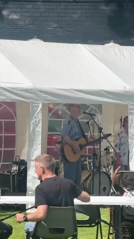 A wedding reception is being held outdoors under a white tent. A band is playing on a stage inside the tent. A male singer is playing an acoustic guitar and singing into a microphone. Another male musician is standing next to him, and a drum set is visible behind them. Several people are seated at tables in front of the stage, and some children are dancing on the grass in the foreground. The reception appears to be taking place on a sunny day, with trees and grass visible in the background.
