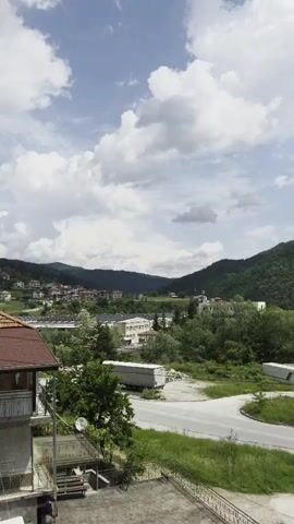 The video captures a static, elevated view of a scenic town, likely Rudozem, Bulgaria, nestled in a valley surrounded by lush green, tree-covered mountains. The sky is partly cloudy with large white clouds against a blue background, indicating daytime.

In the foreground, the corner of a building with a red-tiled roof and a balcony is visible on the left. Below it, a white satellite dish is mounted on a lower structure. A winding asphalt road traverses the middle ground, with green grassy areas on either side. A white car drives briefly from left to right on this road. Further back, various buildings, including residential houses with red roofs and larger, possibly commercial or industrial structures, are scattered amongst abundant trees. A large white truck trailer is parked on the side of the road in the mid-distance. Another larger building with a white dome, possibly a mosque, is visible further back against the foothills. The background is dominated by a series of rolling, forested mountains under the bright sky. No people or visible text are present in the video.