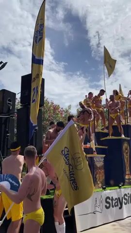 A group of men in yellow speedos and soccer-themed attire are on a decorated float. The float features large speakers, a tiered platform, and flags with the text "Stay Sitges Accommodation." Some men are dancing and holding drinks, while others wave flags. The background shows a blue sky with clouds and trees. A soccer ball is visible on the float. The overall atmosphere appears to be a festive parade or event.