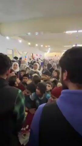 The video shows a large group of children gathered indoors, many looking toward the person filming or toward someone speaking in front of them. The children are of various ages and dressed in casual clothing, with some wearing head coverings. A woman with a white head covering and a light-colored vest with some kind of insignia can be seen carrying a sign with what appears to be a drawing or symbol on it. The environment is a large indoor space with bright overhead lighting, and some decorations such as balloons and banners are visible. People appear to be moving around, and there is an overall sense of excitement and anticipation. The camera perspective is from within the crowd, looking towards the front.