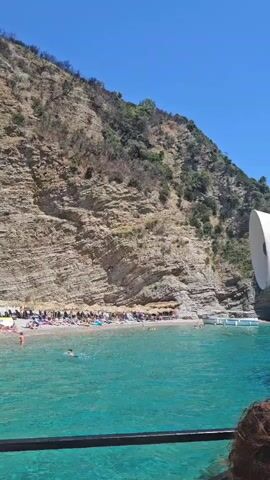 A clear, sunny day at a crowded beach nestled at the base of a steep, rocky cliff. The turquoise water is calm, with several people swimming and wading near the shore. The beach is lined with rows of lounge chairs and umbrellas, indicating it is a popular tourist spot. People are sunbathing, relaxing, and enjoying the water. The cliff face is textured with layers of rock and sparse vegetation. The video appears to be shot from a boat or a dock, with a railing in the foreground.