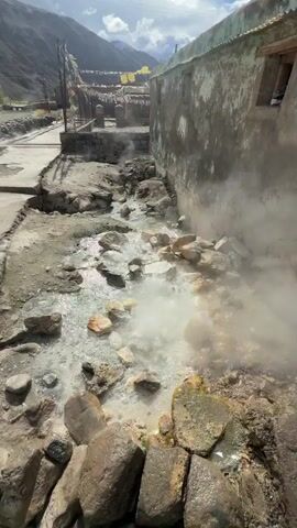 The video shows a narrow, rocky stream with steam rising from it, indicating hot springs. The stream is bordered by stone walls and a concrete path. In the background, there are buildings and steep, rocky mountains. Some prayer flags are visible hanging from a structure. The camera pans across the scene, capturing the natural, rugged environment.
