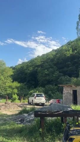 The video begins with a full shot of a white SUV parked on a dirt path, facing away from the camera. The SUV is positioned in front of a partially constructed stone building with a wooden roof. Behind the building, a steep, tree-covered hillside dominates the background, with a clear blue sky dotted with white clouds above. To the left of the SUV, the terrain is uneven and appears to be a construction area, with piles of dirt and some construction materials visible. The camera pans slowly upwards, focusing on the dense green foliage of the hillside and the expansive blue sky. As the video progresses, the camera continues to pan upwards, revealing more of the natural landscape. The SUV remains visible in the lower portion of the frame. The scene appears to be an outdoor rural setting on a sunny day. The video concludes with a wide shot of the landscape, emphasizing the natural beauty of the forested hillside under a bright sky.