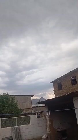 This video captures an outdoor scene under a cloudy sky. The foreground features a low-lying wall constructed from white cinder blocks, topped with a white mesh fence. Beyond the fence, a lush green tree stands to the left, with a smaller, sparser tree to its right. Behind the trees, a building with a corrugated metal roof is visible, appearing somewhat weathered. To the right of this building, another structure is partially seen, its roofline extending towards the right edge of the frame. This second building has a textured, straw-like material on its upper walls and visible roof beams. In the distance, mountain peaks are silhouetted against the sky.

The sky is filled with a dense layer of gray and white clouds, suggesting an overcast or potentially approaching rain. The lighting is muted, indicative of either early morning, late afternoon, or a generally dim, cloudy day. No people are visible in the frame. The overall impression is a rural or semi-rural setting, likely residential, with a focus on the sky and the structures in the mid-ground. There is no visible text in the media file. The muted colors and heavy cloud cover could evoke a sense of calmness or anticipation of weather changes.