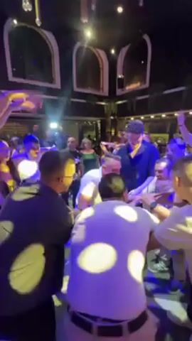 The video shows a group of men dancing in a circle at what appears to be a party. They are mostly dressed in white shirts and the lighting is dark with blue and yellow highlights. The environment looks like a club or party venue, with decorations hanging from the ceiling.