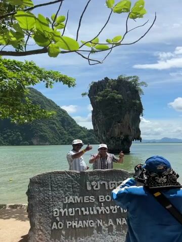 Two men stand behind a large stone marker that reads "JAMES BOND ISLAND" in English and Thai. The man on the left wears a white and blue striped shirt and a white baseball cap. The man on the right wears a patterned shirt and a white baseball cap. Both men are smiling. In the background, a tall, slender rock formation rises out of the water, topped with trees. The water is a calm, greenish-blue. The sky is blue with scattered white clouds. Green mountains are visible in the distance on the left. The image is taken from a slightly low angle, with a branch of a green leafy tree hanging down from the top left corner. A person's arm and shoulder, wearing a blue and white jacket, are visible in the lower right corner, holding up a phone to take the picture.