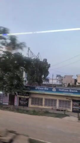 The video captures a street scene with a blurred, moving perspective, suggesting it was filmed from a vehicle. In the foreground, a dirt road is visible, with a motorcycle carrying two people moving across the frame. Behind them, a building with yellow and blue signage is present, one sign indicating "Indian Bank". Further back, a pedestrian overpass with stairs is visible, and beyond that, a white building with domes, resembling a mosque or temple. The sky is overcast. The scene appears to be in an urban or semi-urban environment in India, given the presence of the Indian Bank and the style of the architecture. The overall lighting suggests it is either early morning or late evening.