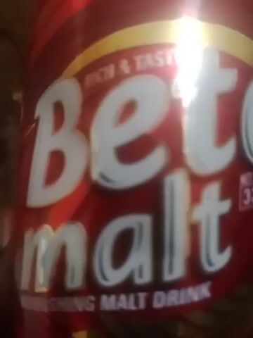 The video shows a close-up of the label of a beverage can. The label is red with white lettering. The word "Bere" is prominently displayed in large white letters, with the top line of text "RICH & TASTY" above it. Below "Bere", the word "malt" is partially visible in smaller white letters. The bottom of the label reads "NOURISHING MALT DRINK" in even smaller text. The lighting suggests the can is illuminated by sunlight, creating some glare and highlights on the text.