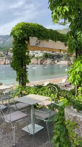 The camera is static, positioned at a slight upward angle, providing a view from what appears to be an outdoor cafe or restaurant. The foreground features a square table with four chairs, all rendered in a light green hue, complemented by lush greenery and vines that frame the scene. Above the table, an awning with the word "Nikito" inscribed in stylized script provides shade.

Beyond the cafe, a picturesque coastal landscape unfolds. A sandy beach lines the edge of a calm, turquoise bay. Several small boats are anchored in the water, some with red buoys nearby. In the distance, a rocky cliff topped with houses and vegetation forms the backdrop. The sky above is partly cloudy, contributing to the serene atmosphere. The overall scene evokes a sense of tranquility and relaxation.