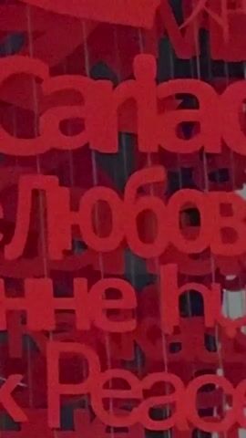 The video shows a large, red, heart-shaped sculpture suspended from the ceiling in a modern, spacious indoor environment. The sculpture is made up of many smaller words and letters, also in red, which appear to be in various languages. The environment appears to be an airport or a large public transportation hub, with high ceilings, large windows, and signage visible in the background. The floor is highly reflective, and the lighting is bright and even. There are a few people walking in the background, and escalators are also visible. The camera slowly zooms out, revealing the scale of the sculpture and its surroundings.