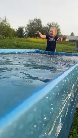 A young girl with blonde pigtails, wearing a dark t-shirt with a colorful design, stands in an inflatable swimming pool filled with clear blue water. She is positioned in the center of the pool, with the water reaching her mid-torso. The pool is rectangular with blue inflatable sides. The background shows a grassy area with trees and vegetation. In the distance, a house and a trampoline are visible. The girl is smiling and actively splashing water with her hands, creating ripples and splashes in the pool. The lighting suggests it is daytime, possibly late afternoon or early evening.