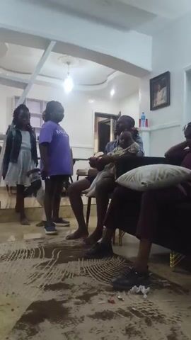 A medium shot captures a family scene indoors. A man is seated on a chair, holding a toddler on his lap, appearing to be watching something. He is wearing a gray shirt and dark shorts. The toddler is in a patterned dress and white socks. A woman stands to the left, wearing a purple t-shirt with "MILANO DI ROUGE" printed on it and dark shorts. Another person is partially visible on the right, wearing a maroon outfit. The room has light-colored walls and a patterned rug. There's a door visible in the background, and a framed picture hangs on the wall. The man appears focused, while the toddler is sitting calmly.