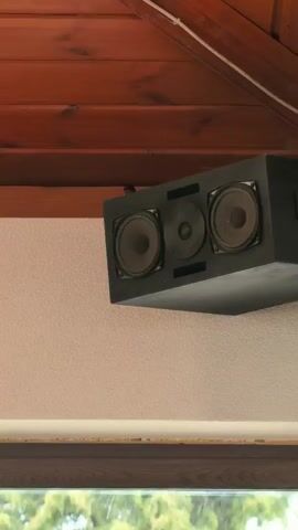 The camera pans up from a speaker to a wood plank ceiling. The speaker is black and rectangular with three visible speakers and is attached to a wall and the ceiling. The ceiling is made of narrow wood planks that are stained reddish-brown. A window frame is visible below the speaker, and light can be seen shining through the window.