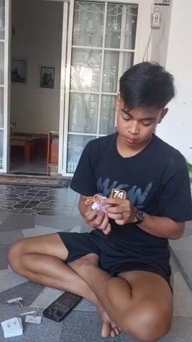 A young man in a black t-shirt and black shorts sits cross-legged on a tiled floor, working with a small object in his hands. He is focused on his task, his head bowed slightly as he manipulates the items. Scattered around him on the floor are a few small papers, some with numbers written on them, and a smartphone. Behind him, a sliding glass door opens to an outdoor area, with a glimpse of furniture inside the building. The scene appears to be in a residential setting, possibly a porch or patio. The man's hands are actively engaged with what appears to be paper and rubber bands, suggesting he is crafting or assembling something.